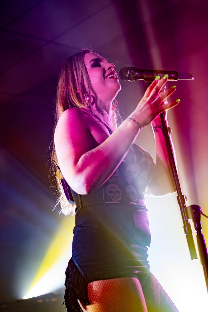 Jessica Barnes; Blissful Photography   Devilskin   Vocalist Jennie Skulander   Mount Maunganui 15th April 2023;I love the lighting/light beams behind Jennie Skulander in this image, right down to her bright fingernails and red lipstick while singing a melody in one of their songs.