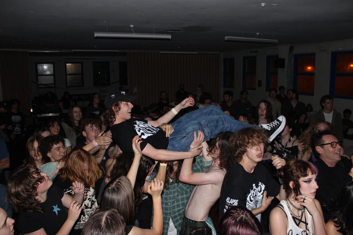 Mars Cook;Jack Crowdsurfing;This photo was taken at an all ages gig during a performance by local Tāmaki Makaurau band Ratso. The red lights outside are from a fire truck that called in the middle of the show. The photo is taken after we were all let back in after evacuating to continue the show so people were pretty hyped. I jumped onto the stage to take it because I'm a bit short. My favourite details are the hands. It was a pretty rough pit but the hands in this shot look graceful and relaxed even though theyre helping carry a person.